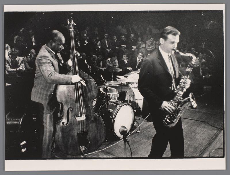 The image captures a live jazz performance featuring two prominent musicians: one playing the double bass and another on tenor saxophone. The setting appears to be an indoor concert hall with an engaged audience in the background, suggesting this was likely a formal event or showcase. Both performers are dressed in suits typical of mid-20th-century jazz ensembles, indicating that the photograph is historical, possibly from around 1959 as suggested by references like "Oscar Pettiford, Kenny Clarke en Stan Getz" and Ed van der Elsken's involvement.

The musician with the double bass has a focused expression on his face, demonstrating concentration or passion for playing. The saxophonist is looking slightly to his left, possibly at another performer or part of the arrangement not visible in this frame. Their instruments are well-maintained and polished, reflecting their professionalism.

There's no discernible text within the image itself that provides additional context beyond these 