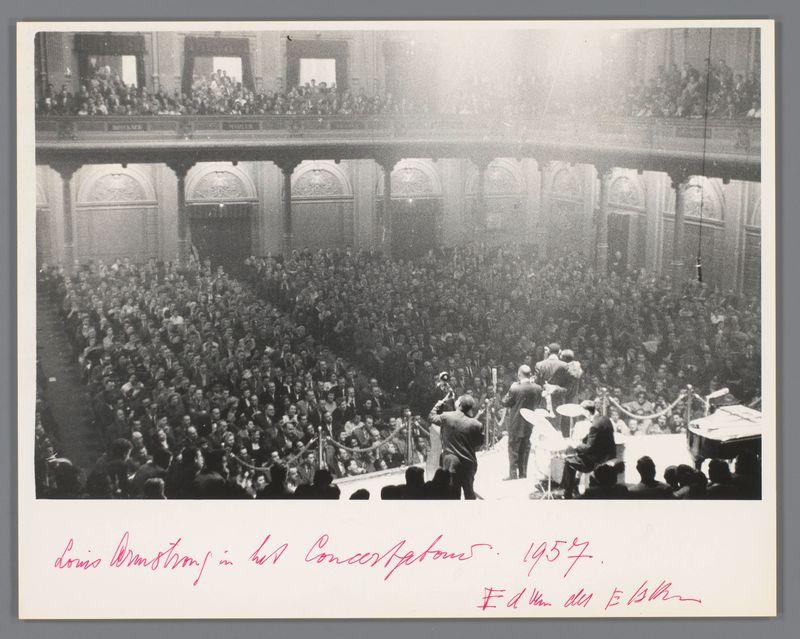 The image depicts a concert taking place in an opulent hall with multiple levels of balconies filled with spectators. A large crowd is gathered, suggesting the event's popularity and significance. At the forefront on stage are musicians, including Louis Armstrong and his All Stars band members, actively engaged in performing. The audience appears attentive and absorbed by the music. Handwritten text at the bottom indicates that this was a concert performance of Louis Armstrong in 1957.