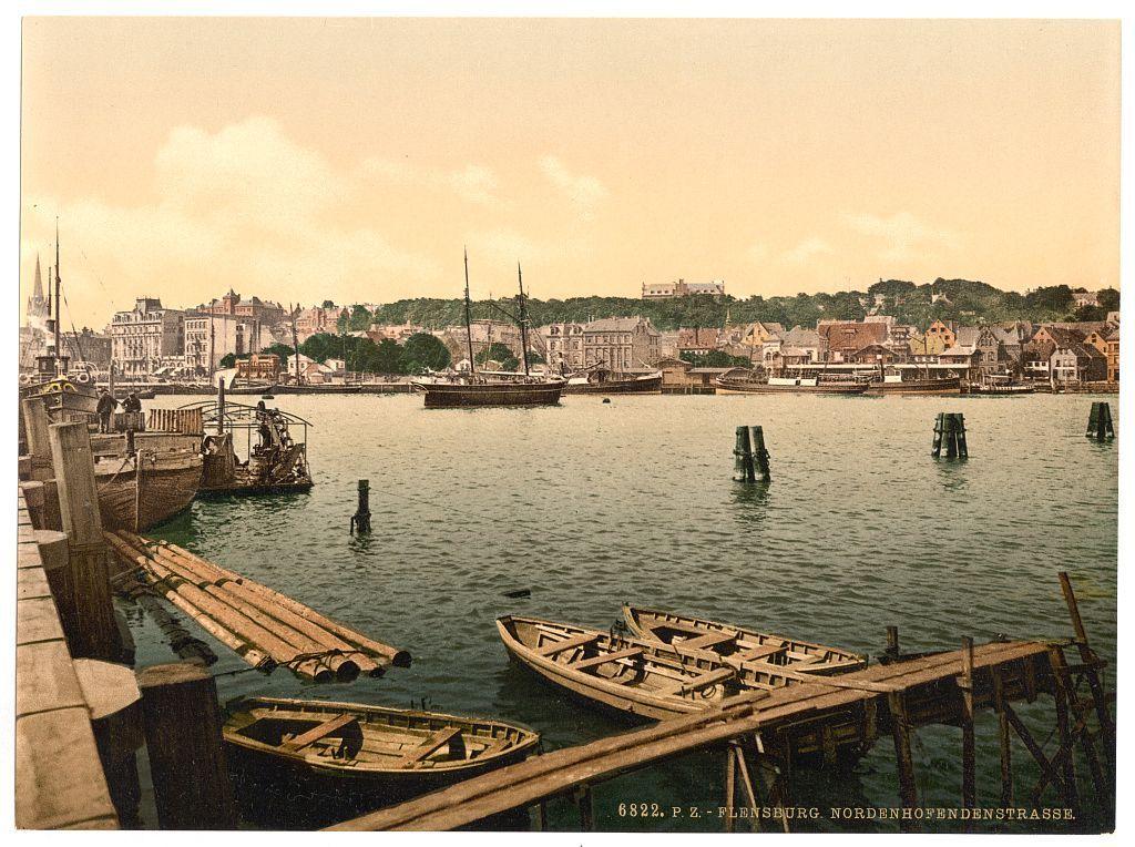 This image depicts a scenic waterfront view with multiple wooden boats moored in calm waters. In the background, there is a bustling harbor filled with larger vessels docked along piers and jetties. The architecture of buildings across the water suggests an urban setting, possibly indicative of early to mid-20th century design. There's noticeable activity as people can be seen on some boats and near docks, implying daily commerce or transportation functions at play.

The sky is overcast with a yellowish hue that may suggest either dawn or dusk lighting conditions. The overall composition evokes the ambiance of maritime life in a European coastal town during an era where such waterfront activities were integral to local livelihoods.