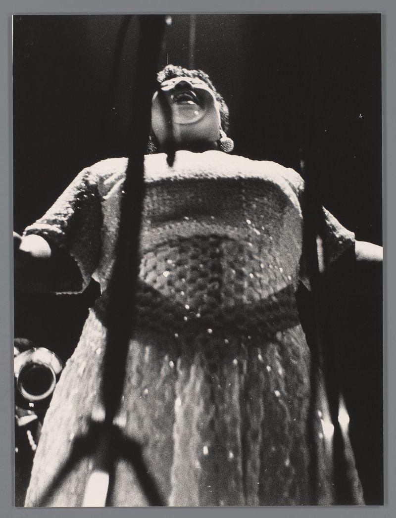 A black and white photograph captures a moment from what appears to be a concert performance. A woman, possibly the singer or performer, is captured in mid-action with her mouth wide open as if she's singing passionately into a microphone that sits on a stand beside her. She wears an oversized dress adorned with intricate patterns, suggesting it may have been chosen for its dramatic impact during performances. The background fades to blackness, focusing all attention on the illuminated figure and hinting at stage lighting used in live music events. A blurred outline of what could be part of musical instruments or possibly audience members is visible around her silhouette, contributing to a lively ambiance. There's no discernible text within this image that indicates its specific context beyond being captured during a performance likely associated with Louis Armstrong given the mention of his name and reference to Amsterdam in the provided source information.