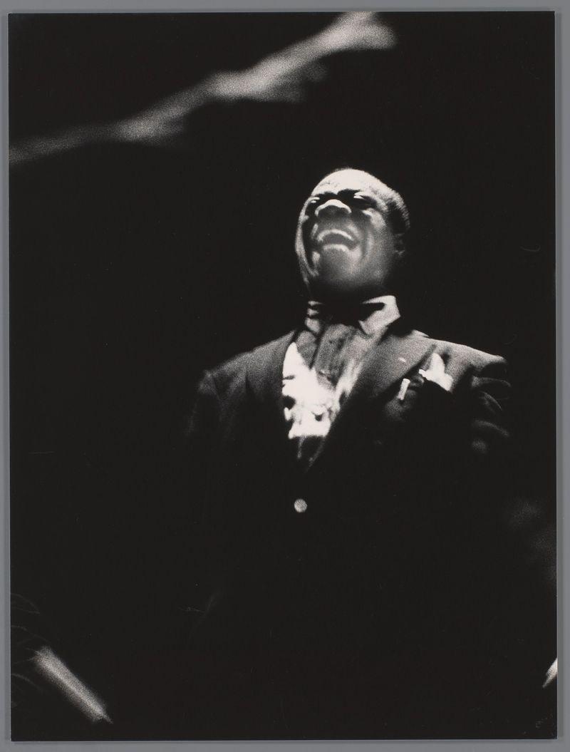 The image is a black and white photograph featuring an individual who appears to be in mid-performance, likely singing or speaking with great passion. The person's mouth is wide open as if caught during a powerful vocal expression, eyes are closed tightly, possibly indicating emotional intensity or concentration on the music.

They are wearing formal attire, including what looks like a dark suit jacket and white shirt with an ornate patterned cravat tied around their neck. There is no discernible background detail that indicates location; it's primarily focused on capturing this expressive moment of performance. The surrounding darkness emphasizes the subject's face and upper body.

This photograph was taken by Louis Armstrong in het Concertgebouw, Amsterdam according to the provided link. It represents a significant cultural icon from 1955 captured during an intimate concert setting which showcases his emotive expression as he performs live on stage for Ed van der Elsken, as indic
