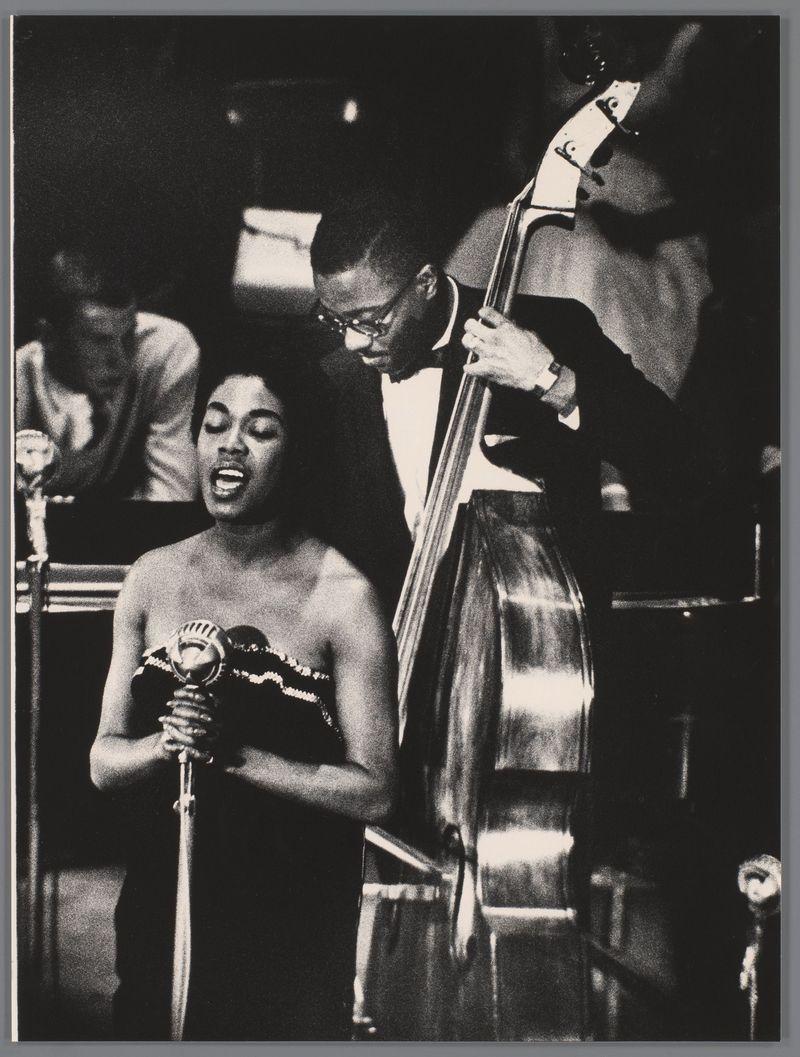 The image is a black and white photograph depicting two musicians on stage during what appears to be a jazz performance. On the left, there's an individual singing into a microphone, wearing a strapless dress with decorative embellishments around the neckline. The person has their eyes closed and mouth open as if in mid-song. To the right stands another musician playing a double bass, attentively looking down at his instrument while holding its neck piece between his fingers.

The setting is indicative of an intimate live music venue or concert hall ambiance, with focused lighting on the performers contrasting against a darker background that includes other musicians and equipment such as additional microphones. The style of clothing worn by both individuals suggests this photo may be from mid-20th century jazz performances. Notably, in the bottom right corner is a caption credit attributing the image to Ed van der Elsken with information on its source provided via hyperlinks linking