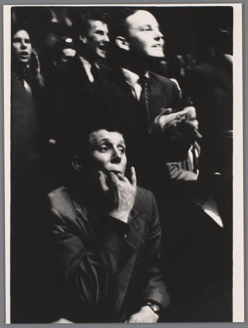 The image captures a moment from what appears to be an indoor concert performance, as suggested by the presence of individuals in formal and semi-formal attire. The focus is on four people seated closely together: two men standing behind them with expressive facial features suggesting engagement or excitement about something happening off-camera; one man sitting in front with his hand thoughtfully placed under his chin, looking upward towards an unseen point above him that could be the stage where Lionel Hampton's concert might have been taking place. The black and white photograph has a grainy texture indicative of older film stock used during mid-20th-century photography.

The men are dressed in dark suits with ties or collared shirts, which is typical attire for such occasions. One man on the right stands out as he looks upward with an open mouth expression, possibly singing along to the music or reacting vocally to a performance element. The setting seems informal yet lively, cha