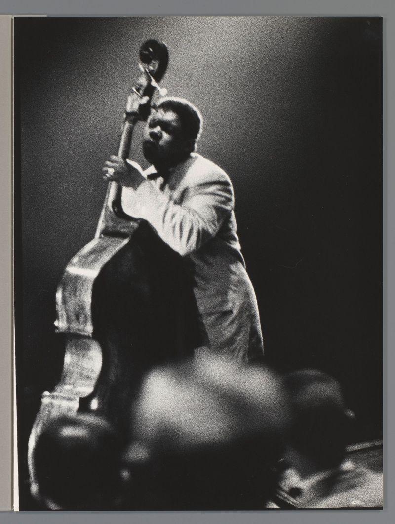 This black and white photograph captures a moment of intense musical performance. A man, likely a bassist given the instrument he's holding which appears to be an upright double bass or contrabass fiddle, is in mid-motion with his head bent downwards towards the bow near its tip as if playing a note. His facial expression suggests concentration and passion for music.

He wears what seems to be casual attire - possibly jeans rolled up at the bottom of their leg openings leading down into dark socks, suggesting an informal setting or personal comfort during performance. The subject's posture is slightly hunched over with his left arm raised higher than his right, indicating that he may have been in a standing position and now has adjusted to play.

The backdrop fades from darkness towards light near the figure of the musician, which focuses attention on him as the central element of this composition. There are no discernible features or objects around him; it's an empty space void of