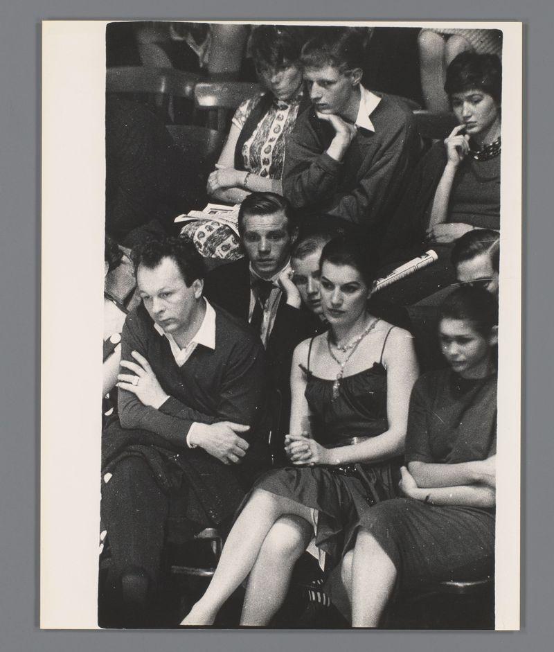 A black and white photograph capturing a candid moment at what appears to be a concert, showing various individuals seated in an audience. From left to right: A man with his arm around another's shoulder, both focused on the event; behind them sits a couple closely together, deeply attentive; next is a young woman seemingly lost in thought, clutching her chin; she’s followed by two more women and men who are similarly absorbed in the performance before them. Their attire suggests an era of classic elegance with dark suits and dresses featuring decorative elements like pearls or beads. The setting indicates an indoor venue, likely a concert hall given the caption reference to Sarah Vaughan's Amsterdam Concertgebouw event.