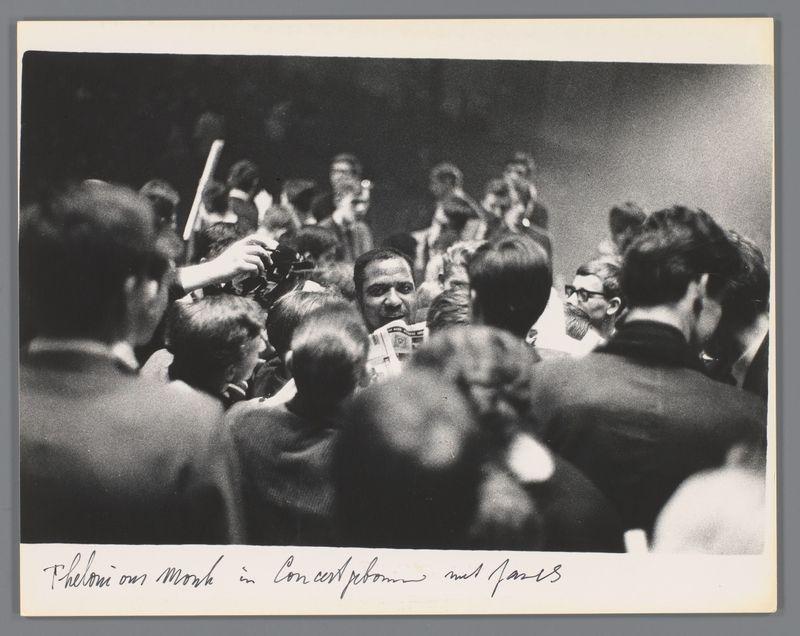 The image captures a bustling scene filled with numerous individuals converging closely together, indicative of an event or gathering where fans have gathered around the central figure. The photograph is in black and white, emphasizing shades ranging from deep blacks to stark whites, creating high contrast that draws attention directly toward the middle ground populated by people.

Amongst this crowd, a person holding up what appears to be a newspaper stands out due to their positioning near the center of focus; they are captured with only part of them visible. Their gaze is directed towards something or someone not within view, possibly engaging with another attendee whose head can be seen directly in front of theirs.

The surrounding individuals display various postures and expressions that suggest attentiveness, curiosity, or engagement with each other or the central figure's activities. The absence of color conveys a timeless quality to this moment, potentially situating it wit