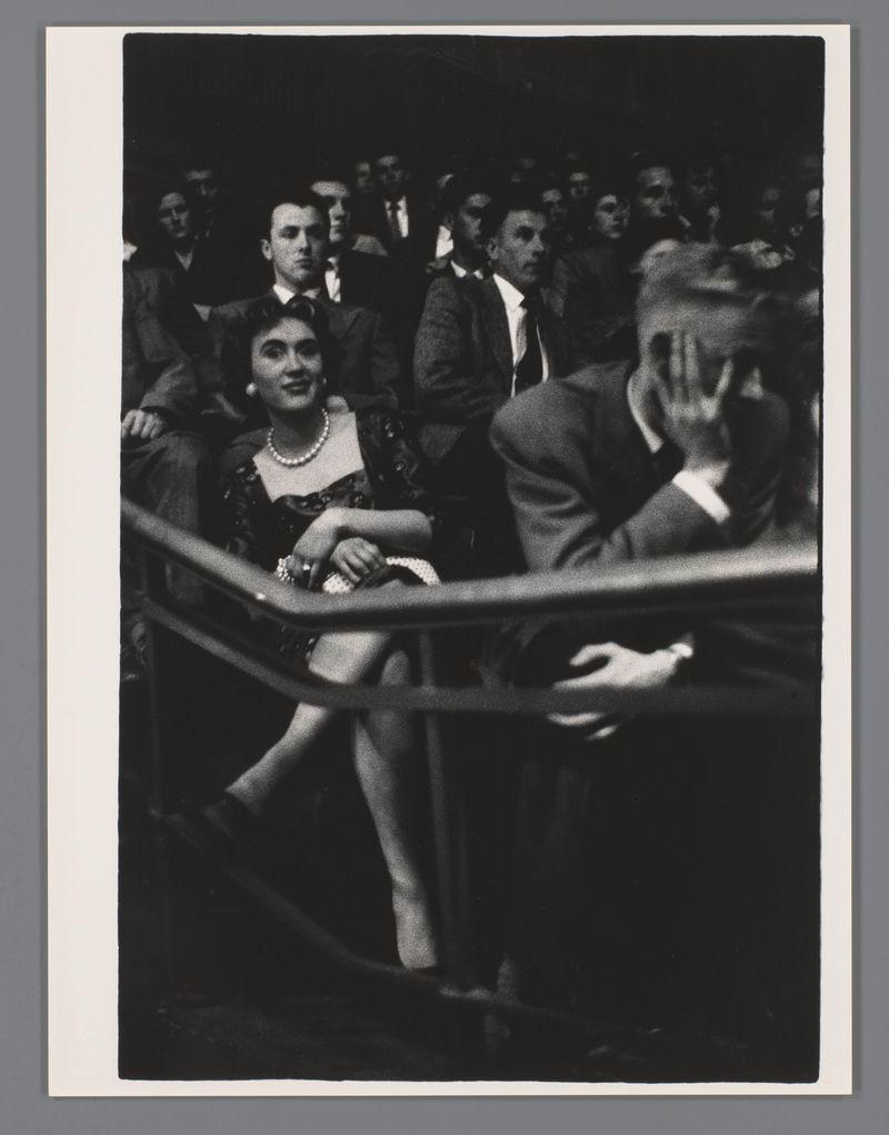 The image is a black and white photograph featuring an audience at what appears to be a concert or public event. In the foreground, two individuals are seated on either side of each other; both dressed in formal attire suggesting a level of sophistication typical for such events. The woman wears a dark dress with pearls around her neck, indicating evening wear. Her gaze is directed off-camera, and she has a slight smile.

Adjacent to them sits an individual whose face is obscured by their hand, possibly shielding eyes from bright stage lights or avoiding being photographed themselves. In the background, multiple figures are visible in various states of engagement; some look on attentively while others engage with each other or seem distracted.

The lighting focuses more intensely on those at the front and fades into darkness towards the edges of the frame, creating a sense of depth and isolating the audience from their surroundings. The overall mood is somewhat somber yet dignified