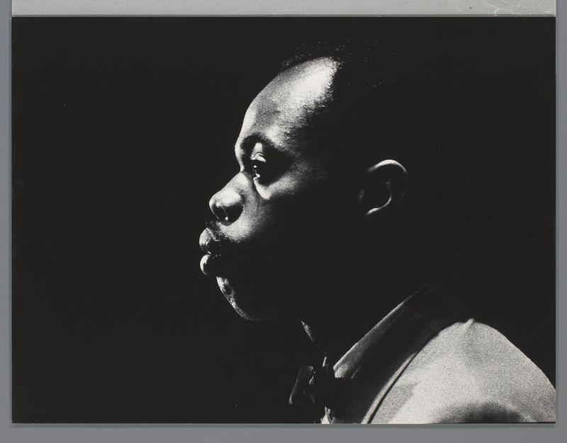 The image is a black and white photograph of an individual captured in profile. The person appears to be engaged with something, possibly playing or speaking into a mouthpiece that's held near their lips, suggesting the act of blowing music such as jazz saxophone. This could indicate that they're either practicing or performing live. There are notable shadows across parts of the face and clothing due to the strong lighting from one side, which accentuates textures like hair and fabric folds.

The background is dark, focusing all attention on the subject who stands out with lighter shades where their skin catches light against a contrasting black backdrop likely meant for dramatic effect or artistic expression. The individual's attire appears formal but not overly so, potentially suggesting this could be an informal studio session rather than an official photo shoot.

There are no discernible colors due to it being monochrome; the range of tones varies from white highlights and grey