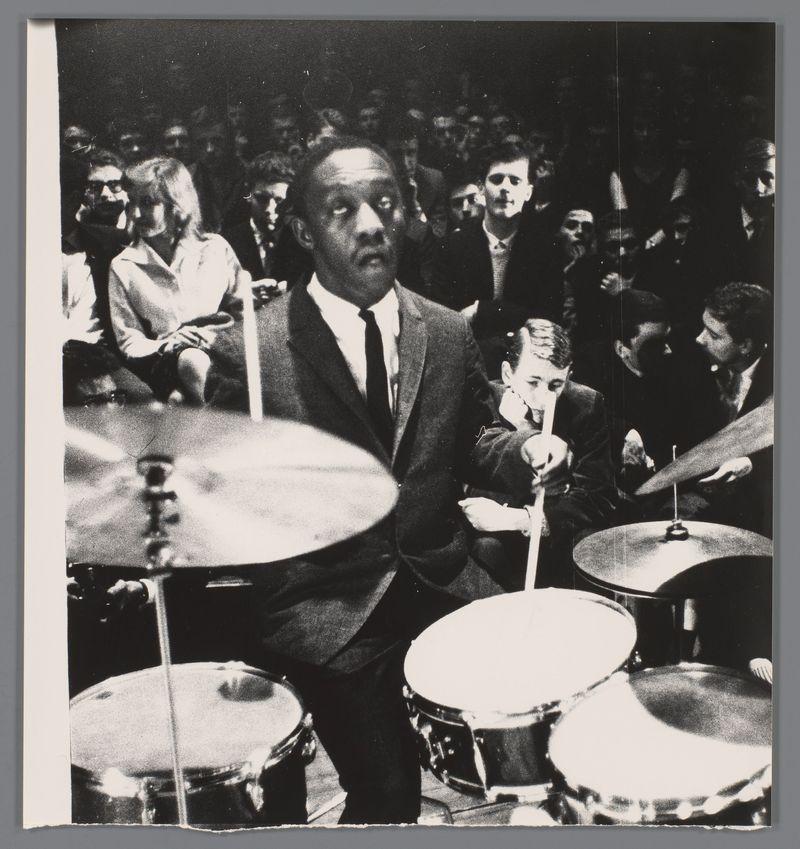 The image captures a moment from what appears to be an energetic jazz performance, featuring drummer Art Blakey in the foreground. He is dressed formally with a suit and tie, intensely focused on his drum set which includes at least two cymbals and several drums of various sizes. His posture suggests he's either playing or has just finished a spirited beat.

The background shows a group of engaged spectators seated closely together around him, their attention directed towards the performance. The audience members display varied expressions—some are intently watching Blakey while others appear to be conversing among themselves. Their attire is more casual compared to his formal wear.

This black and white photo carries an air of vintage appeal, likely from a 1958 concert as indicated by the provided source link which credits Ed van der Elsken for capturing this scene in Amsterdam's Concertgebouw. The overall composition conveys both the passion of live jazz music and its communal ex