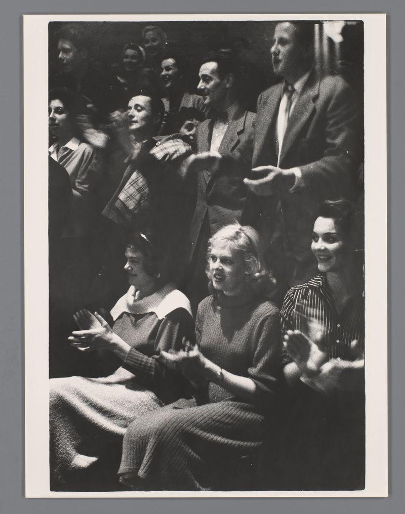 The image is a black and white photograph capturing an audience seated closely together, likely in the context of a concert or performance. The individuals appear to be engaged with what they are watching or listening to; several people have their hands raised as if applauding or clapping along. Their expressions range from focused attention to enjoyment.

In terms of attire, it suggests mid-20th-century fashion: some women wear dresses and cardigans while men don suits with ties. The photo's composition centers on the collective group rather than individual faces, giving a sense of communal experience.

The background is mostly indistinct due to depth-of-field effects but appears to be an indoor venue typical for performances or public gatherings during that era. No specific details about the performers are discernible from this angle and perspective within the crowd.

This image seems captured by Ed van der Elsken, known for his work in Amsterdam around 1956, as referenced in t