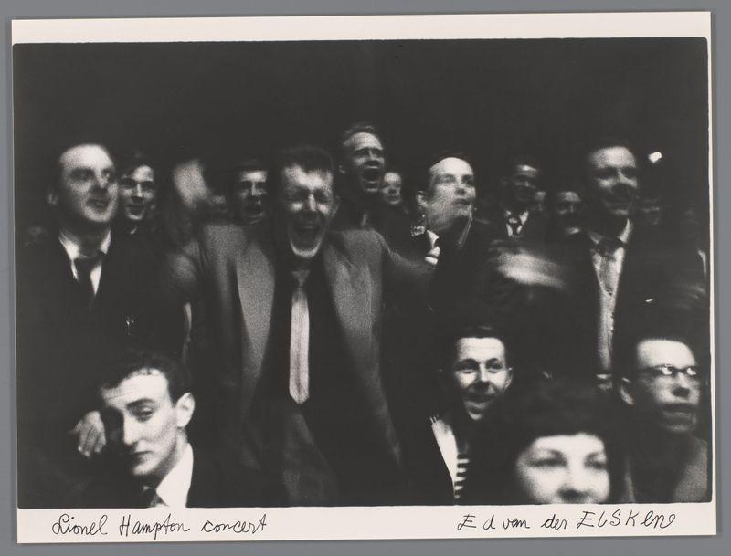The image is a black and white photograph capturing the moment at what appears to be a concert. It's taken from an elevated angle, showing several people in close proximity, likely audience members or perhaps performers, with varying expressions of excitement or intensity on their faces.

In the foreground, there are two prominent figures: one man is captured mid-laugh or shout with his mouth wide open and eyes squinted shut, suggesting a moment of high energy. Next to him sits another individual looking directly at the camera; he appears pensive or contemplative, gazing downwards towards his left side.

Behind these individuals, there's a crowd that seems engaged in the event. The lighting is stark with strong contrasts between light and shadow on their faces, emphasizing facial expressions but obscuring finer details of features like eyes or ears due to the shadows cast by surrounding figures' heads.

The bottom right corner has handwritten text which reads "E d van der Elske" 