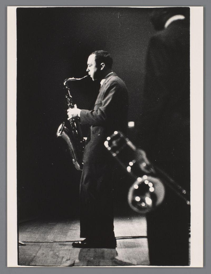 This black and white photograph captures a tenor saxophonist from the J.J. Johnson Quintet performing live on stage in Amsterdam's Concertgebouw, 1957. The image is taken by renowned Dutch photographer Ed van der Elsken, as indicated by the source link provided.

The musician is captured mid-performance, playing his saxophone with a focused intensity that suggests he is deeply immersed in his musical expression. His attire appears to be formal, suitable for the period and setting of performance photography. The lighting highlights him against a dark background, emphasizing the spotlight effect often used during live performances, which draws attention directly onto the performer.

The composition includes another musician partially visible on the right side, indicating that this was likely part of an ensemble or band setup typical in jazz concerts from that era. The grainy texture and high contrast are characteristic of film photography from the mid-20th century, adding to the hist