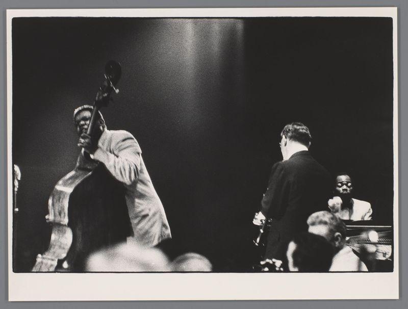 The image is a black and white photograph capturing a moment from what appears to be a live jazz performance. In the foreground, there's a musician prominently featured holding a bass guitar with an expressive posture that suggests deep concentration or emotion during his playing. His attire includes a light-colored shirt which contrasts against the dark background of the stage.

In the middle ground, another individual is visible; he seems to be standing beside a piano and possibly engaging in conversation or listening intently to the performance as indicated by his stance with one hand on the body for support. Behind him stands an audience member who appears engaged with what's happening onstage but not directly involved in it.

In the background, we can see part of another musician seated at a keyboard instrument which could be a piano or organ and is partially obscured from view due to perspective. The lighting focuses attention primarily on the musicians while leaving much of 