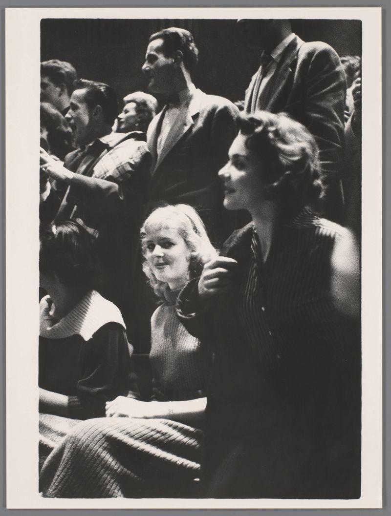 The image is a black and white photograph capturing a candid moment of people in what appears to be an audience setting. There are at least four individuals visible, with the focus on two women sitting close together; one slightly behind the other, both facing towards their left where something off-camera seems to have caught their attention. The woman closest to us has blonde hair and is wearing a light-colored sweater or shawl draped around her shoulders. She appears contemplative or possibly concerned as she looks away from whatever event they are attending.

The second woman in the foreground wears a dark, patterned dress with short sleeves and a collar; her head is turned towards the first woman, suggesting an interaction between them. The person directly behind these two women seems to be leaning forward slightly, engaging more intensely with the ongoing activity or performance outside of view.

In the background, there are silhouettes and partial profiles of other people who