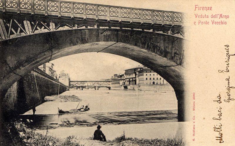 The image shows a vintage postcard featuring the Arno River and the Ponte Vecchio in Florence, Italy. The photo captures an arch bridge spanning over the river with ornate truss work on its underside. Below is another view of the bridge from beneath it; two individuals are visible: one sitting by the water's edge and another standing at a distance near the bridge.

In the background, there is historic architecture along the riverbank, including what appears to be a large building or palace with multiple stories and classical design elements. The sky is overcast, casting diffused light across the scene which gives it an aged appearance typical of historical photographs from early 20th-century postcards. Red cursive handwriting on the top right corner reads "Firenze" followed by Italian phrases translating to 'Florence,' along with references to Arno and Ponte Vecchio.

The bottom left side bears a red stamp that says "G. Modiano e Co - Milano," indicating it is from Milan, Italy in 
