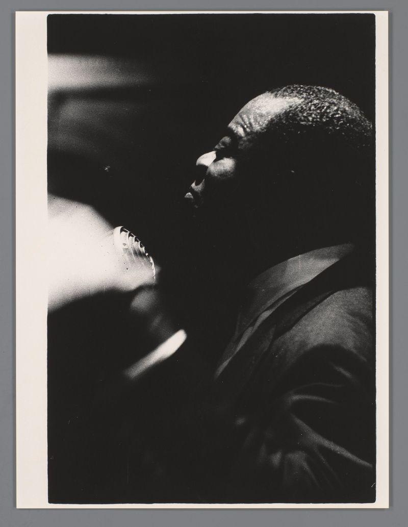 The image shows a man playing the trumpet with intense focus and passion. His facial expression is one of deep concentration, as he appears to be deeply immersed in his performance. The photograph captures him from an angle where we can see part of his profile against a dark background that accentuates his silhouette. There are no colors visible since it's a black-and-white image; however, the contrast between light and shadow adds depth and drama to the composition.

The trumpet is held close to his mouth, with fingers placed on its valves, indicating an active playing moment. The instrument itself has a reflective surface that catches some of the ambient light in the room where he performs. His attire appears casual yet professional—a common choice for jazz musicians during performances—adding authenticity and context to the image.

The background is blurred, drawing attention entirely to the musician's performance without any distractions. This style of photography emphasizes no