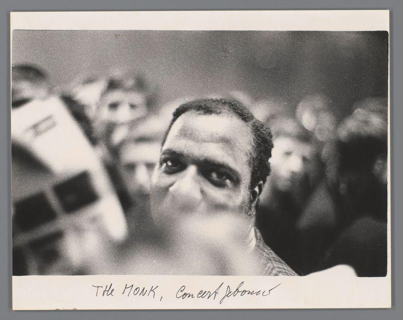 The image is a black and white photograph featuring the face of an individual in close-up, with blurred figures surrounding them. The person appears to be Thelonious Monk based on the text annotation provided at the bottom which reads "The Monk," suggesting that this could indeed be a portrait or depiction associated with him. There's additional handwritten script indicating "Concertgebouw" and what seems like his signature in cursive, hinting at an association with Amsterdam Concert Hall.

This photograph is part of Ed van der Elsken's collection, as indicated by the source link provided which references Portret van Thelonious Monk bij een optreden in het Concertgebouw. The year mentioned suggests it was taken in 1961 during a concert event at this location. Van der Elsken is known for his work capturing various aspects of life and culture, particularly within urban environments.

The image presents an intimate yet candid portrayal with the focus on Monk's facial expression which 