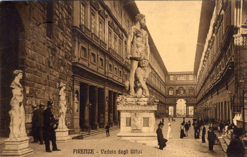 The image depicts a historical black and white photograph of an outdoor courtyard in Florence, Italy. In the center stands a large stone statue depicting two figures: one appears to be a muscular man with a lion-like creature on his back, while another figure seems to be a satyr or mythological being beneath them. The statue is placed within a rectangular pedestal adorned with various sculptural decorations.

The courtyard surrounding this central sculpture features classical architecture, including detailed stone facades and arched windows typical of Renaissance-era buildings. People are visible in the scene; some walk along the pathways while others stand near the statues or engage in conversation.

In the background, through an open archway, one can see more architectural structures extending into a distant courtyard area, suggesting this location is part of a larger complex. The image also contains text at the bottom that reads "FIRENZE - Veduta degli Uffizi," which translates 