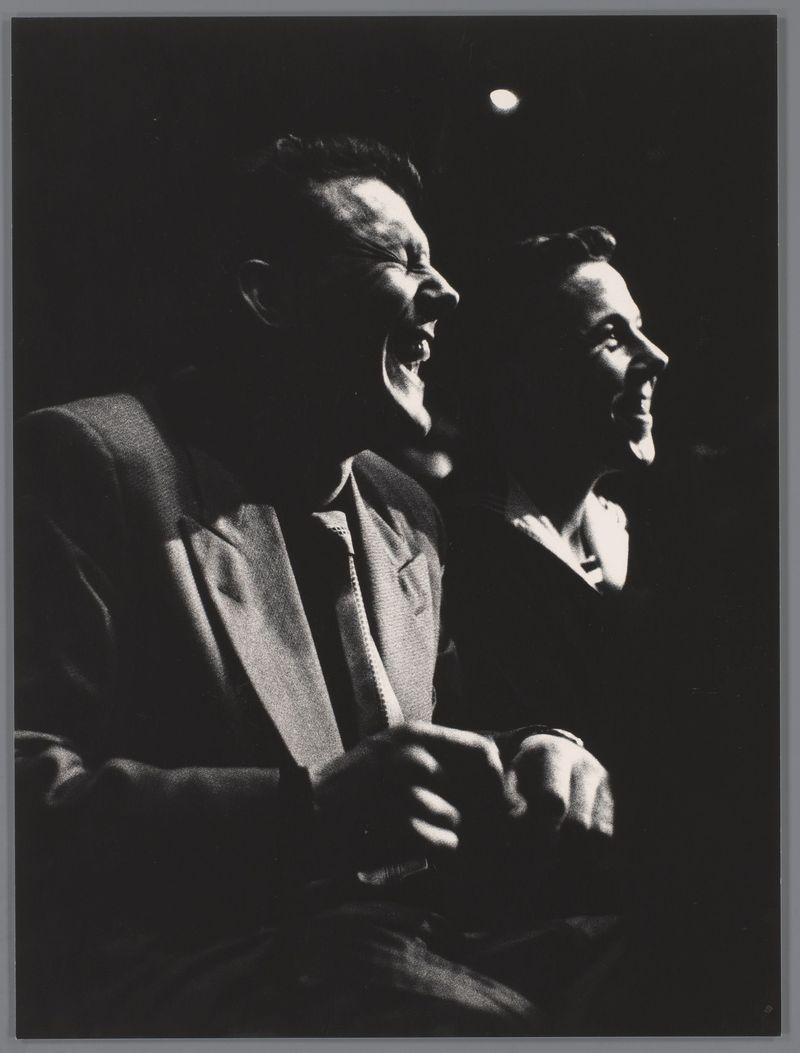 The image is a black and white photograph capturing two individuals seated side by side in what appears to be an auditorium setting, likely during or after a performance. The individual on the left has their mouth open as if laughing or cheering enthusiastically, with visible lines of light suggesting bright stage lighting above them. This person's hair stands up around the head due to movement, possibly from laughter or excitement.

The second individual is slightly turned towards the first but also appears engaged and joyful, indicated by a wide-open expression that suggests they are reacting positively to an event out of frame. Both individuals wear dark clothing with discernible texture, which could be indicative of formal attire suitable for such events. The background is mostly obscured in darkness, focusing attention on their expressions and reactions.

The photograph's lighting highlights the faces and upper bodies of both subjects while keeping much of the surrounding envi