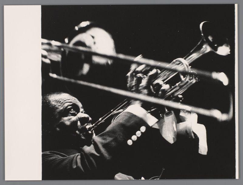 The image is a black and white photograph depicting two musicians in the midst of a performance. The musician on the left, with his face partly obscured by shadow, appears to be playing a trumpet or trombone-like brass instrument, as suggested by its cylindrical shape and mouthpiece proximity. His right hand is visible near the valves, indicating active manipulation during play.

The musician on the right is captured in motion, possibly blowing into an unseen horn like instrument given his open mouth expression and blurred posture indicative of movement. The photograph conveys a sense of action and musical engagement between both performers with their instruments held up towards each other as if interacting harmoniously through music.

No discernible background elements are present; the focus is entirely on capturing the moment of performance, highlighting the musicians' concentration and energy in what appears to be an intimate setting.