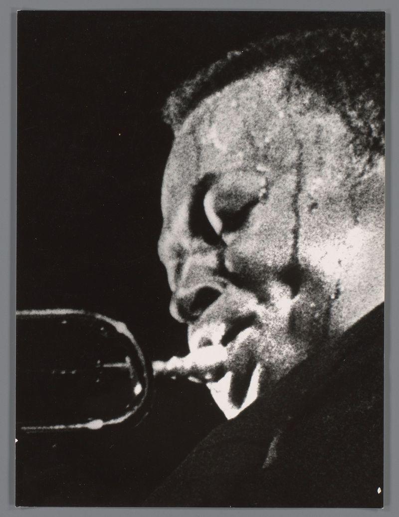 The image is a black and white photograph of an individual playing the trumpet. The focus is on the musician's face as they blow into the instrument, capturing detailed expressions that suggest concentration or emotion in their performance. The background is dark and indistinct, emphasizing the subject. There are no discernible colors due to the monochromatic nature of the image. No additional context or setting details can be provided from this frame alone.