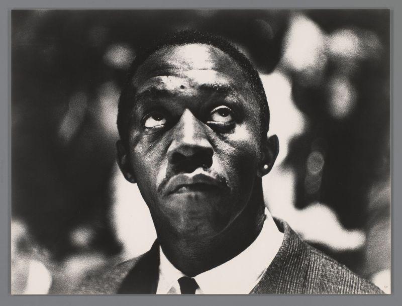 The image is a black and white photograph of a man, with the focus on his face which occupies most of the frame. His expression is contemplative or serious as he gazes slightly to one side away from the camera's perspective. He appears middle-aged with defined cheekbones, deep-set eyes, and visible wrinkles around them suggesting age and perhaps an active lifestyle. The lighting accentuates these features sharply against his skin tone.

He wears a suit jacket over what seems to be a light-colored dress shirt paired with a dark tie. His ears are adorned with earrings, which stand out prominently due to the high contrast of the image. There is significant shadowing in the background, indicating that this photograph was likely taken during an event or gathering as there appears to be indistinct figures and lights blurred behind him.

The overall composition suggests a formal setting, possibly a concert given some context clues like "in het Concertgebouw" which could hint at being Amst