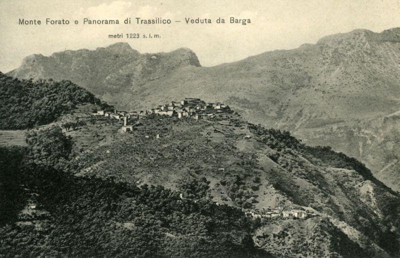 The image is a black and white photograph depicting a rugged, mountainous landscape. The scene features several clustered buildings atop a hill or mountaintop, which suggests the location could be a village situated in a remote area of Italy as indicated by text on the photo mentioning "Monte Forato e Panorama di Trassilico - Veduta da Barga," and additional details about altitude ("metri 1223 s. l. m."). The terrain is steep, with sparse vegetation covering rocky outcrops and gentle slopes leading up to where the buildings are positioned. The overall impression conveyed by this historical photograph is of a serene yet isolated community within a scenic mountainous region.