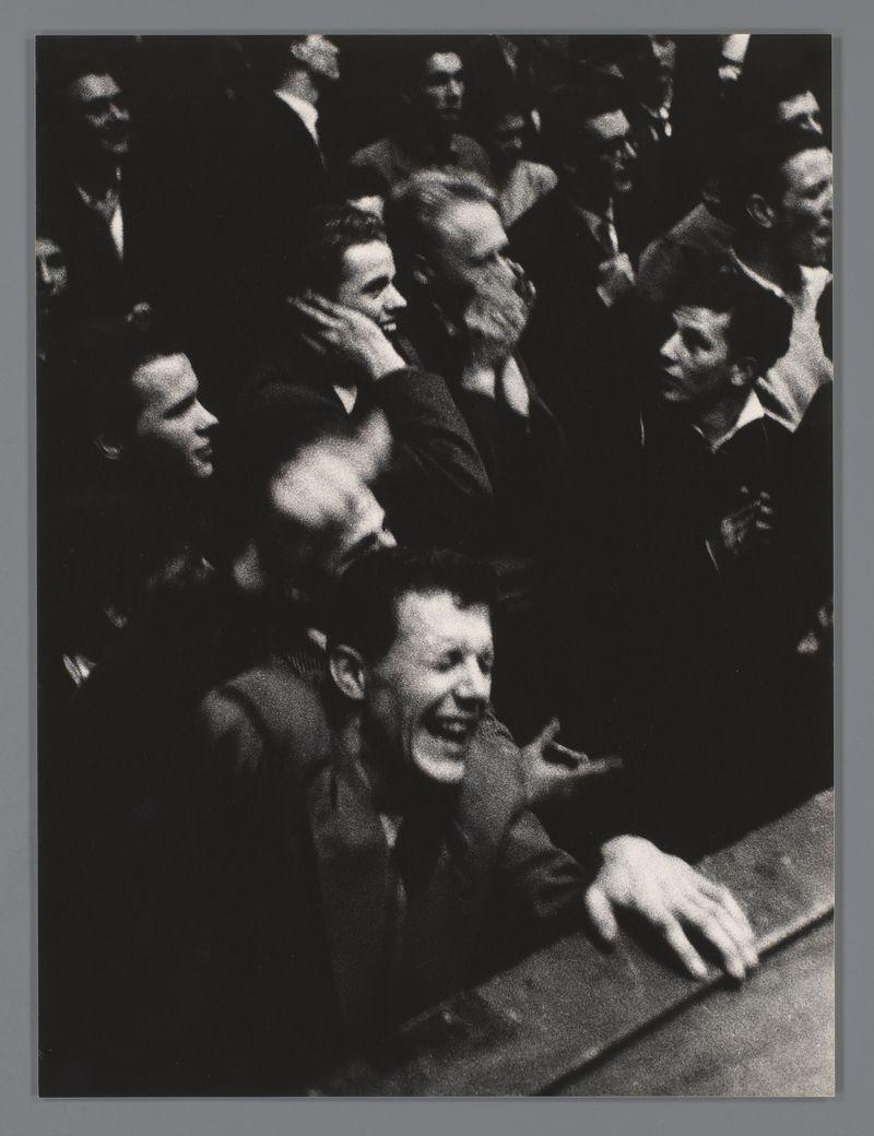 This black and white photograph captures a moment from what appears to be a concert in the Concertgebouw, Amsterdam. It is taken by Ed van der Elsken during Lionel Hampton's performance in 1956. The image prominently features an individual smiling broadly with their hand on a table or barrier, seemingly engaging enthusiastically with the audience. Surrounding this central figure are various individuals who seem to be part of the crowd, some raising their arms and others clapping. Their expressions range from joyous laughter to attentive interest, indicating a lively atmosphere typical of concert settings. The lighting is dramatic, highlighting faces while casting shadows that contribute to the energetic mood conveyed by the scene.