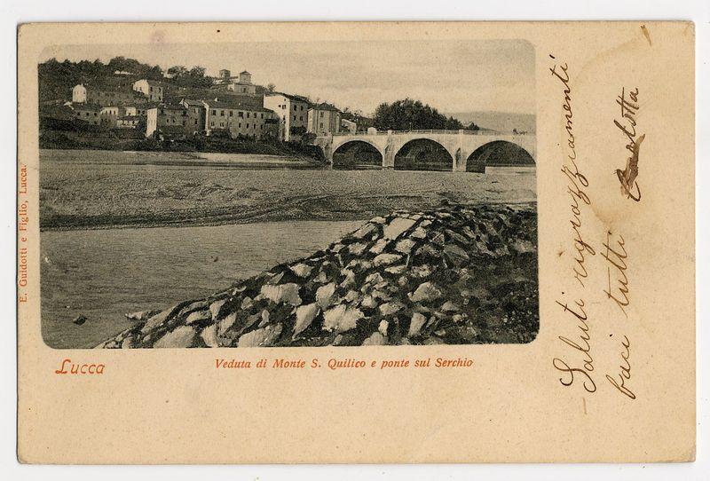 The image is a vintage postcard showing an old photograph of the Monte S. Quirico and its bridge on the Serchio River in Lucca, Italy. The black-and-white photo depicts a riverside scene with waves crashing against rocks near the riverbank. Across the water lies a town built into hillside terraces; buildings are clustered together, leading up to larger structures at higher elevations. A notable feature is an arched bridge spanning multiple arches across the river. Handwritten text in cursive script adorns one side of the postcard, possibly indicating it was sent as correspondence with some additional note or message included. The bottom left corner bears a red stamp-like imprint reading "Lucca," while at the top right, there is another handwritten phrase suggesting personal sentiment associated with the photo's viewing.