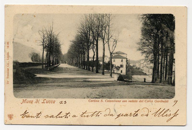 The image is a sepia-toned vintage postcard featuring an old European street scene. The card's title "Mura di Lucca 2" suggests it depicts the walls of Lucca, Italy, specifically location number 23 on Corso Garibaldi. It shows a tree-lined avenue leading towards distant hills or mountains shrouded in mist.

The left side of the postcard includes handwritten text that reads "Ganti saluti a tutti da parte gli sti" and is signed with an unclear name ending with "-t." The handwriting appears personal, possibly from someone who visited this location. In contrast to the handwritten note, printed on the right-hand side are details in Italian: "Cortina S. Colombano con veduta del Corso Garibaldi 9," which translates to "Cortina San Colombano with a view of Corso Garibaldi." The text also includes an identifier number and location information that suggests it is part of a larger collection.

The card's borders are worn, indicating age or frequent handling. There's visible wear at the top ri