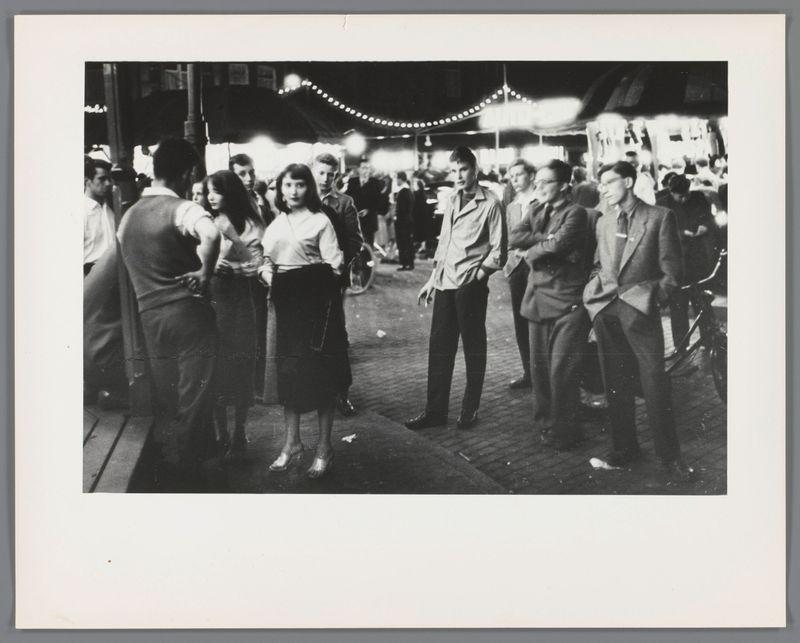 This black and white photograph captures a bustling street scene with numerous individuals gathered in what appears to be an outdoor market or fair. The crowd is diverse, featuring men and women dressed in attire indicative of mid-20th century fashion—a mix of casual suits for the males and various dresses or skirts for the females. Some people are engaged in conversation while others seem absorbed in activities around them.

The street is adorned with string lights, adding a festive ambiance to the setting. The background reveals additional market stalls, hinting at food vendors or goods being sold, though specific details of these stalls are not discernible from this perspective.

Notably, one woman stands out as she holds what seems to be two cigarettes in her hand—a detail that could suggest social norms regarding smoking during that era. There is a bicycle parked on the right side, suggesting accessibility for cyclists or perhaps another mode of transportation common at the ti