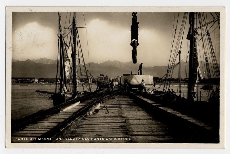 The image is a black and white photograph depicting an industrial maritime scene. It shows a large dock extending into calm waters with mountains faintly visible in the background, suggesting it might be taken during late afternoon or early evening as indicated by the soft lighting.

On the left side of the dock, there's a sailing ship moored alongside, its sails furled and rigging prominent against the sky. The deck is cluttered with various maritime equipment such as ropes and possibly cargo handling tools, indicating active use for loading or unloading goods.

A significant feature in this image is an industrial crane situated on the right side of the dock's entrance; it appears to be lifting a cylindrical object into place. Two figures are visible near the crane: one standing atop its structure observing the operation, and another by the water's edge possibly overseeing the process or preparing for loading.

The photograph includes text in Italian at the bottom which translat