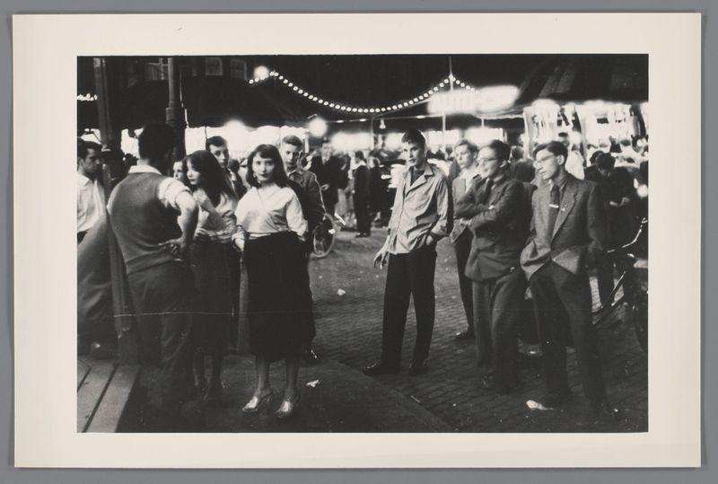 The image is a black and white photograph capturing a lively street scene at night. It appears to be taken in an urban environment, possibly during a fair or festival given the presence of string lights above that suggest a festive atmosphere. A group of people are gathered on what looks like wooden planks laid out as pathways for pedestrians.

In the foreground, there is a woman standing with her back slightly turned towards us; she has long dark hair and wears a light-colored blouse paired with a darker skirt or dress. Surrounding her are men in various states of conversation and movement: some facing each other, possibly engaged in discussion, while others seem to be passing by without interacting directly.

The middle ground shows more individuals moving through the space, dressed in attire that suggests an era from mid-20th century fashion – women wearing dresses or skirts with tops and men sporting suits, jackets, ties, and hats. The crowd is diverse, indicating a public even
