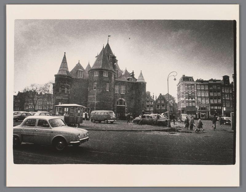 The image depicts a black and white photograph of an urban scene, likely from the mid-20th century. It features Gothic-style architecture with pointed spires and steeples in the foreground, which appears to be an old building possibly used as a town hall or courthouse due to its prominent design and central position.

In front of this structure is a busy street filled with various types of vehicles including cars from different eras—indicating that this photo might capture a transitional period between older automobiles and more modern ones. There's also a vintage Volkswagen bus, which suggests the time frame could be around 1960s to early '70s.

Pedestrians are seen on the street; some appear to be walking while others may be standing or moving about in groups. The clothing of these individuals seems consistent with mid-20th-century fashion—suggesting a cool climate as most people seem dressed for cooler weather, perhaps autumn or spring.

The background includes other buildings