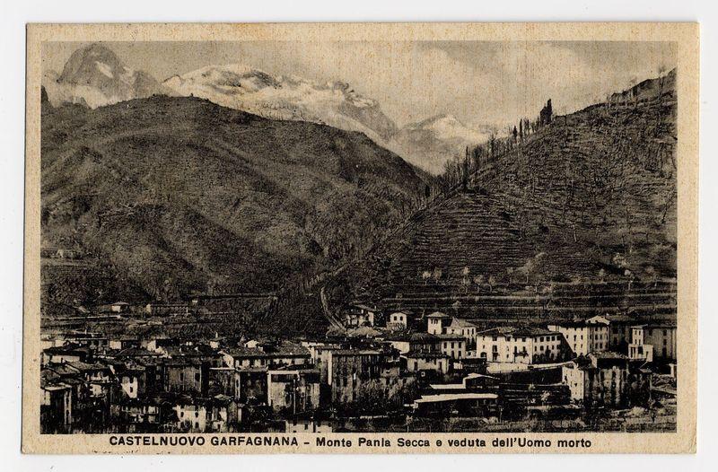 The image shows a vintage black and white photograph of the town Castelnuovo Garfagnana, featuring Monte Pania Secca on its outskirts. The landscape captures rolling hills leading up to snow-capped mountains in the background, indicative of an alpine region. In the foreground is a densely packed urban area with numerous buildings closely constructed together, displaying European architectural styles characteristic of towns nestled within mountainous regions.

The photograph appears aged and shows signs of wear along its edges, suggesting it's quite old or has been used frequently as a postcard. Below the image, there is text in Italian that translates to "CASTELNUOVO GARFAGNANA - Monte Pania Secca e veduta dell'Uomo Morto," which serves as an informative caption identifying both the location and the viewpoint depicted.

The overall composition of this vintage photograph offers a glimpse into historical urban development near natural mountainous terrain, illustrating how towns were 