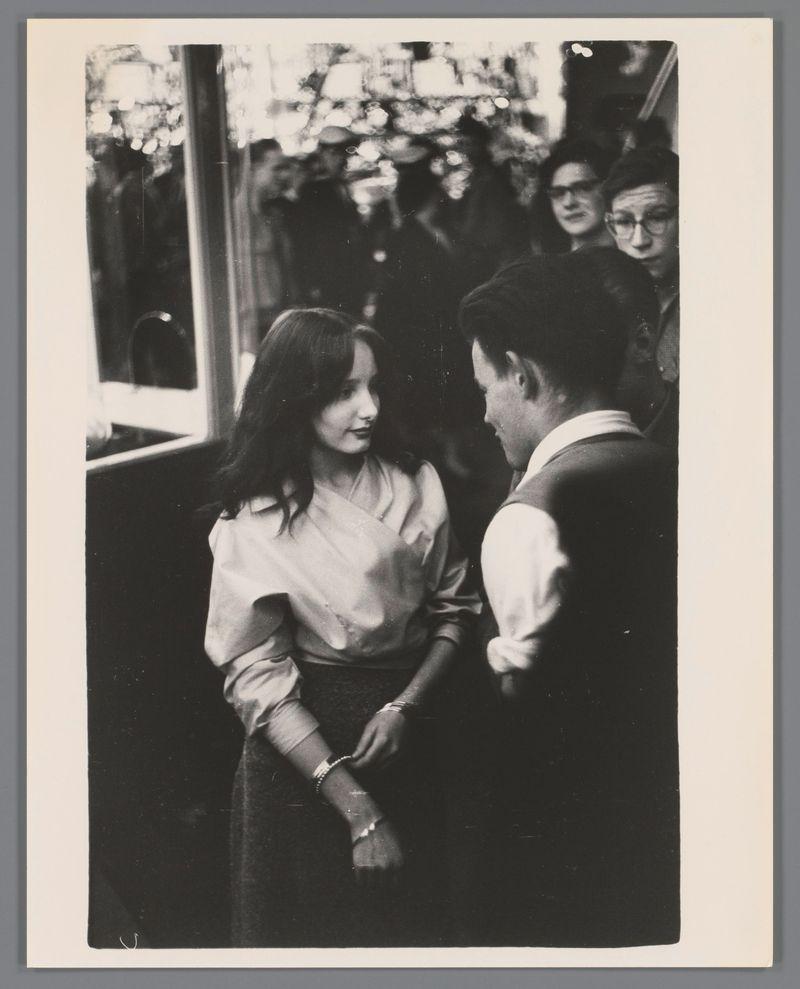 The image is a black and white photograph featuring two people in the foreground who appear to be engaged in conversation. The person on the left, presumed female based on appearance, has long hair and wears what looks like a light-colored dress or blouse with visible buttons down the front, paired with dark pants. She also sports pearl-like bracelets and watches on her wrists.

The individual on the right is dressed more formally; he's wearing a vest over a white shirt and appears to have an open jacket revealing part of his chest. His hair seems combed back neatly.

Behind them stands another person who partially obscures their face, possibly capturing or looking at something not directly related to what we see here. This individual wears glasses and has dark-toned clothing with noticeable patterns on the cuffs that resemble stripes or checks.

The background is quite busy; it's a public space filled with people suggesting an event like a festival. The lights above give off hin