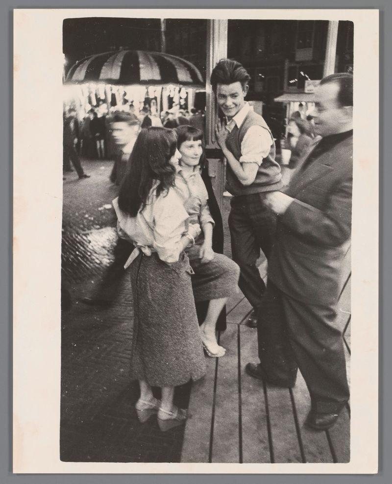 The image is a black and white photograph showing four individuals in what appears to be an outdoor setting, possibly at a fair or amusement park. On the left side of the photo, there are two women standing close together; one woman has her arm around the other's shoulder, suggesting familiarity between them. They both have dark hair, with one wearing a light-colored blouse and darker skirt while the other is in a white top and dark dress. In front of these individuals stands an older man, likely middle-aged or elderly, dressed in casual attire that includes trousers and what seems to be a vest; his expression suggests he might be conversing with them.

To the right, there's another individual who appears to be smoking while engaging in conversation, possibly with one of the women on the left. This person is wearing dark pants, a lighter-colored sweater, and has their arm extended as if gesturing during speech or pointing at something off-camera. The background includes an umbrella-c
