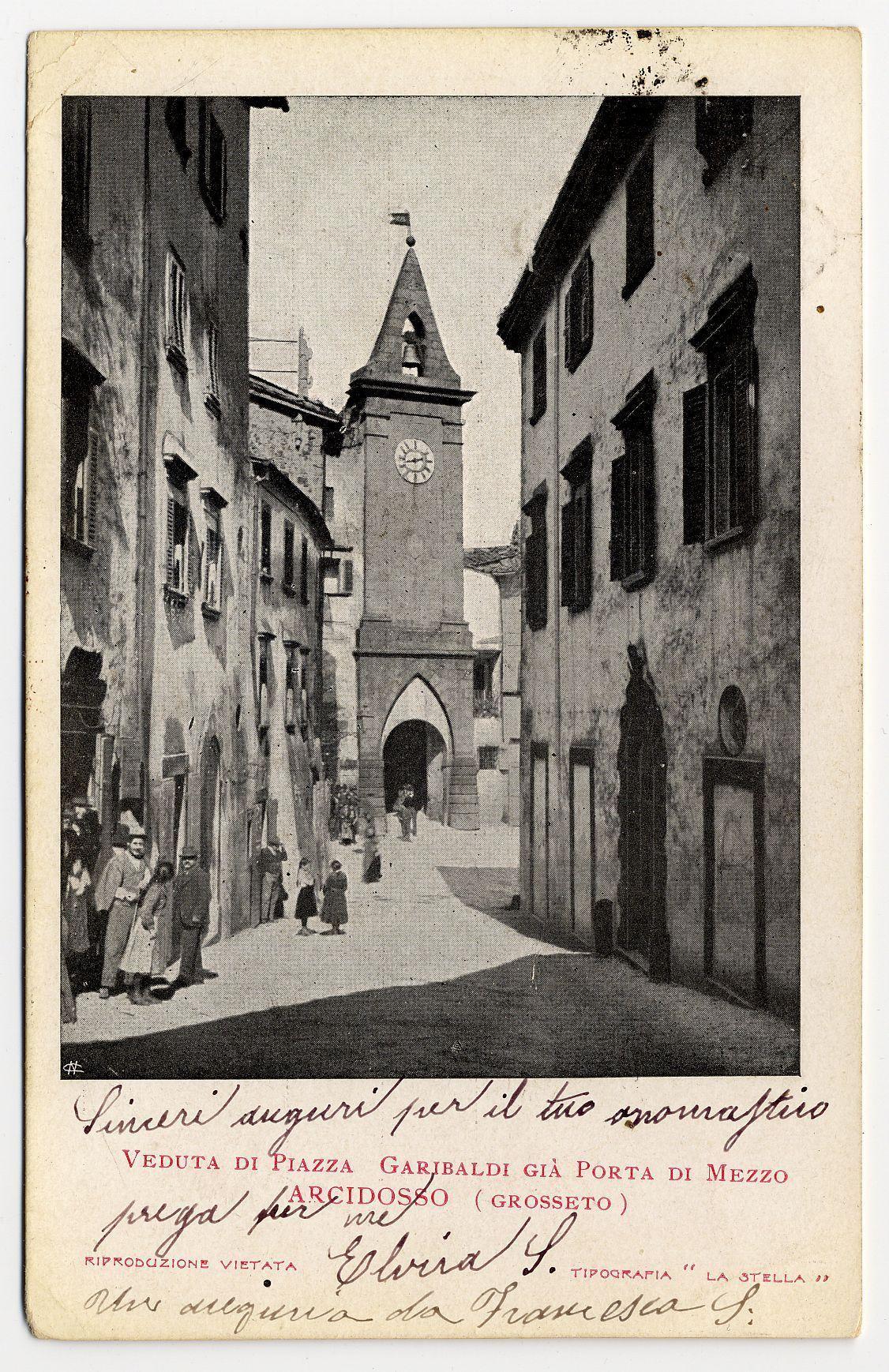 The image is a vintage black and white postcard depicting an old European street leading up to a prominent tower with a clock face, identified as "Porta del Mezzo" in Grosseto. The photograph captures the essence of daily life during that era, showing people dressed in early 20th-century attire walking along the cobblestone path. On either side are traditional buildings with shuttered windows and doors, indicative of historical architecture typical of Italian towns at the time. There's handwritten text on the left-hand side providing context about the image: "Sincere auguri per il tuo anniversario Veduta di Piazza Garibaldi Gia Porta del Mezzo Presso Archidossa (GROSSETO)". The bottom right has additional inscriptions from a reproduction company and signature, suggesting that this postcard was part of an official series or collection.