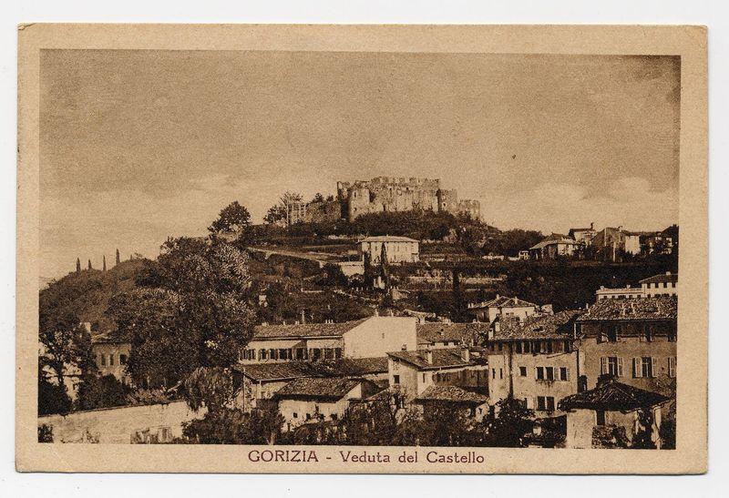 The image depicts an old sepia-toned photograph or postcard of a town with historical architecture, prominently featuring what appears to be a fortress on elevated ground in the background. The foreground shows various buildings with pitched roofs and terracotta tiles that suggest Mediterranean influences. There are trees scattered throughout the landscape, which gently slopes upwards towards the fortress atop a hill.

The text at the bottom indicates "GORIZIA - Veduta del Castello," suggesting that this location is Gorizia, specifically viewed from Castle Hill or within sight of it. The photograph seems to capture an early 20th-century perspective, judging by its monochromatic appearance and historical architecture style.

For further details on a specific image referenced in the alt text (GORIZIA- Veduta DEL CASTELLO), one would need to follow the hyperlink provided for additional information or context about that particular photograph.