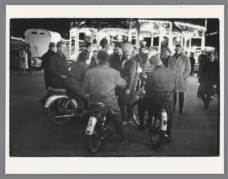 The image captures a group of individuals gathered at night-time, in what appears to be an amusement park or fairground setting. The scene is bustling with activity and includes several people standing around two older motorcycles which are the focal points within this social gathering. These motorbikes have visible kickstands down indicating they might be parked for some time.

Around these central figures, a mix of adults can be seen engaging in various activities; one person appears to be gesturing or pointing towards something out of frame while others look on attentively. The attire suggests it is cold weather as many individuals are dressed warmly in coats and jackets.

In the background, there's an illuminated carousel with its traditional wooden horses and lights creating a festive atmosphere. Adjacent to this attraction stands another large vehicle that looks like a caravan or trailer, labeled "STALLING". This indicates possible commercial activity at the fairground such a