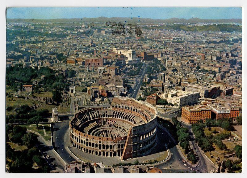 The image displays an aerial view of the Colosseum, a prominent ancient amphitheater located in Rome. The iconic structure is prominently centered and features its characteristic multi-leveled arches which were once used for gladiatorial games and public spectacles. Surrounding this historic landmark are various other architectural structures indicative of Roman antiquity, including buildings with similar stone construction.

The Colosseum sits within a large urban area filled with modern-day developments interspersed among the ancient ruins, showcasing Rome's layered historical narrative. The cityscape extends outward in all directions from the amphitheater, displaying an array of rooftops and streets that wind around it. In the background, there is evidence of green spaces such as trees or parks, providing a contrast to the dense urban fabric.

The colors captured suggest a clear day with ample sunlight casting shadows on some buildings while others are in direct light, enhancing