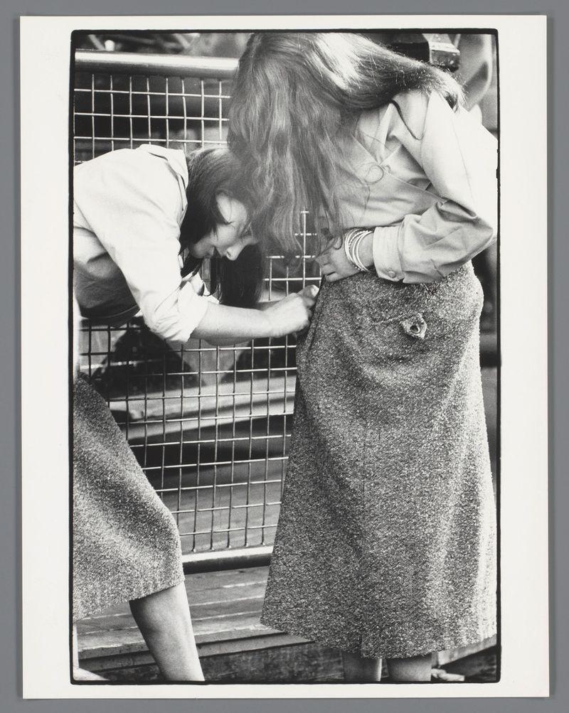 The image depicts two women interacting closely with a wire mesh structure. The woman on the left, wearing a white blouse and skirt, is leaning over to look through or adjust something within the grid pattern of metal bars that form the mesh. She has dark hair tied back and appears focused on her task.

Next to her stands another woman dressed in a textured woolen skirt with buttons down the side seam and what seems like a coat draped around one shoulder. Her light-colored, possibly white blouse is slightly askew as she leans over toward the wire structure. The focus of this second person's attention isn't clear from their posture.

The background suggests an urban setting; however, specific details are indistinct due to the angle and distance at which it was captured. Both individuals display a casual demeanor in what seems like everyday attire appropriate for cooler weather or formal occasions. There is no visible context indicating why they're engaged with this wire mesh structu