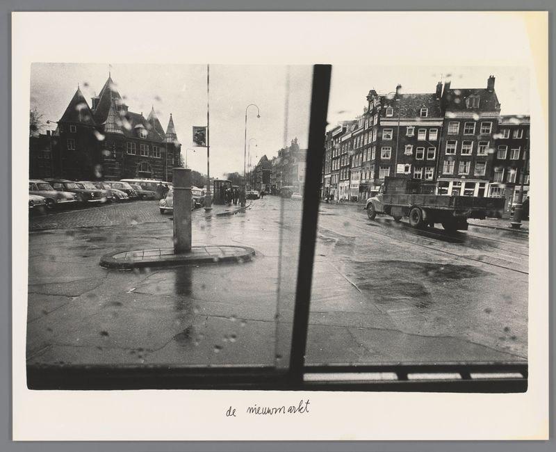 The image is a black and white photograph that captures an urban street scene viewed through the wet glass of what appears to be a window. Raindrops streak across the pane, suggesting recent precipitation or ongoing drizzle. The focus of the shot centers on 'de nieuwmarkt,' likely referring to Nieuwmarkt Square in Amsterdam as indicated by the text at the bottom right.

In this monochrome tableau, we see traditional Dutch architecture with gabled facades lining a wet street that reflects the buildings and vehicles above it. A variety of vintage cars are parked along one side while others traverse the road. Pedestrians can be observed walking on the sidewalks or crossing the streets amidst what appears to be light traffic.

The photograph captures both human activity and stillness, with people dressed in coats indicative of cold weather. The image's composition provides a glimpse into daily life within this European city during its mid-20th-century history, showcasing not only archi
