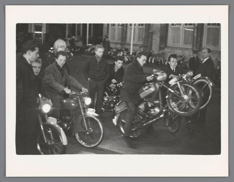 The image is a black and white photograph depicting several young individuals gathered around motorcycles in an urban setting. The photo appears to be from the mid-20th century, judging by the style of clothing worn by the people present as well as their haircuts. There are at least nine visible subjects; some are on bicycles while others stand or sit with a motorcycle.

The men and women displayed varied attire: dark coats, jackets, suits, and trousers dominate the color palette, suggesting cooler weather typical of autumn or winter months in Europe during that era. The motorcycles themselves have classic designs, indicating they may be from an earlier model period rather than contemporary bikes.

In the background, more people can be seen, as well as a row of parked motorbikes which suggests this could be at some kind of social event like a fairground or gathering place in Amsterdam's Nieuwmarkt area. The lighting is dim and seems to emanate from street lamps or interior lights, 
