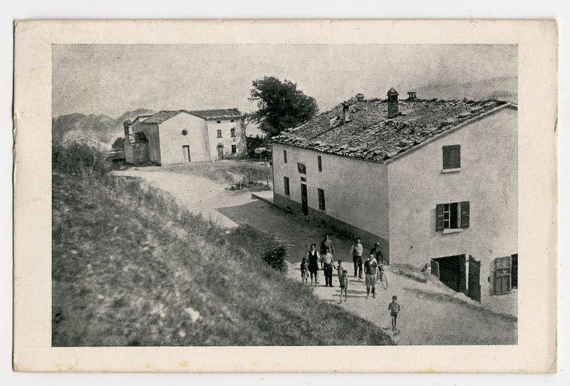 The image depicts a black and white photograph of an older, rustic village scene. It shows several buildings with aged facades, including what appears to be a main house on the right side featuring dark shutters over its windows and a prominent tiled roof adorned with chimneys and vents. To the left stands another building with similar architectural style but less detailed in appearance due to distance or focus.

A group of people is seen walking along a narrow pathway that runs from the bottom center towards these buildings, suggesting a rural setting where pedestrian pathways are directly adjacent to residential areas. The individuals appear dressed for cooler weather and some wear hats. They seem engaged in conversation as they move together down the path.

In the background, rolling hills can be discerned under an overcast sky, which adds to the historical ambiance of the scene. A solitary tree stands on a slope near the top left corner, adding depth and emphasizing the rural s