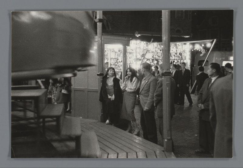 The image captures a bustling scene within an indoor market or fair. A group of individuals is seen engaging with the environment, walking and interacting in what appears to be a lively atmosphere. The grayscale nature of the photograph suggests it may have been taken several decades ago, possibly during the 1950s as indicated by the provided source information.

The people are dressed in attire that reflects mid-20th-century fashion—women wear skirts or dresses and men sport jackets with trousers. Their expressions range from conversation to contemplation, contributing to a sense of everyday life captured at this public event.

The market is filled with various stalls adorned with items for sale; the exact products are indiscernible due to the monochrome palette and focus on people rather than goods. The architecture includes pillars or beams that suggest an industrial-style roof structure common in markets from past decades, adding context to the setting of this photograph.