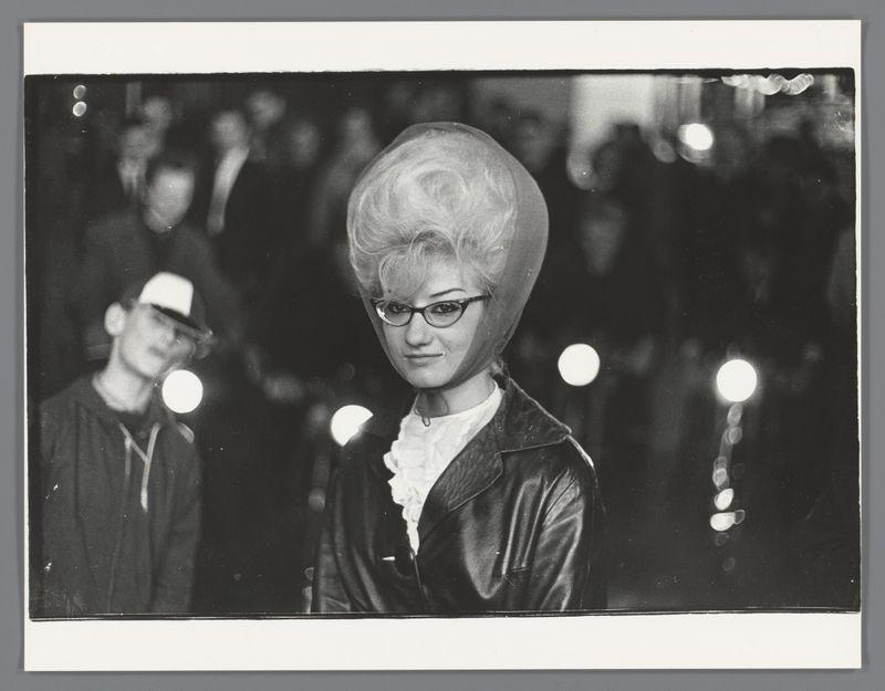 The image is a black and white photograph featuring two individuals. In the foreground, there's an individual with voluminous hair that resembles a beehive style from the 1960s fashion era, wearing glasses, a dark leather jacket over a lighter-colored blouse or dress, which has ruffled shoulders indicative of high necklines popular during that period. The hairstyle and clothing suggest this person may have been attending an event where such styles were fashionable.

In the background, slightly out-of-focus compared to the main subject due to depth of field typically used in photography for emphasizing a portrait, is another individual wearing what appears to be a dark cap or hat with a white visor. This second figure seems to also wear glasses and has their head turned away from the camera, gazing towards something off-frame.

The photograph captures an evening scene suggested by artificial lighting visible as blurred circles that resemble light sources in bokeh effect typical of s
