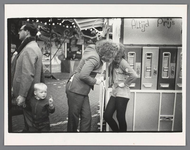 The image depicts a black and white street scene with several individuals engaging in various activities. In the foreground, there is an older man on the left side of the frame wearing glasses and a coat, looking away from the camera towards something out of view. Beside him stands a small child who appears to be gazing at his shoes or possibly examining them closely.

In the middle ground, we see another individual bending down near what seems like a food stand with compartments labeled "Altijd Prijs" (Always Price), suggesting it may offer fixed price items. This person is wearing glasses and has curly hair; they are engaged in an activity involving their hand reaching towards something within one of the compartments.

There's also someone standing opposite to this individual, facing away from the camera, potentially interacting with or observing them.

The background shows a street setting with various objects like vending machines lining what appears to be a market area. The 