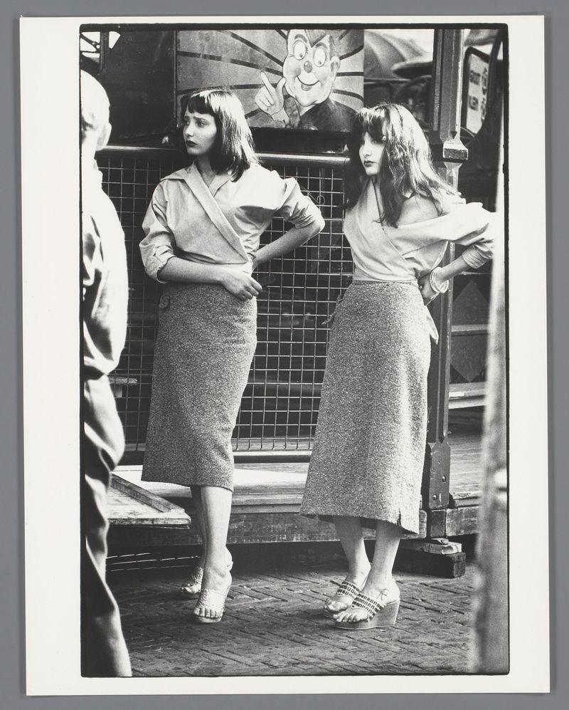 The image is a black and white photograph depicting two women standing side by side on what appears to be the deck of a boat. The woman on the left has short, straight hair with bangs that cover her forehead, dressed in a light-colored blouse tucked into high-waisted trousers, paired with platform sandals adorned with what seem like small flowers or embellishments. She is posing confidently with one hand resting on her hip and turned towards us.

Beside her stands another woman with long wavy hair cascading down the back of her shoulders, wearing a loose-fitting blouse that's slightly untucked over high-waisted trousers. Her footwear consists of patterned open-toe sandals. She too adopts a relaxed pose, hand on her hip and gaze directed away from the camera.

Behind them is an intricate metalwork fence or barrier with geometric patterns, which seems to be part of the boat’s deck design. In front of this structure hangs a large poster featuring cartoonish characters, possibly for co
