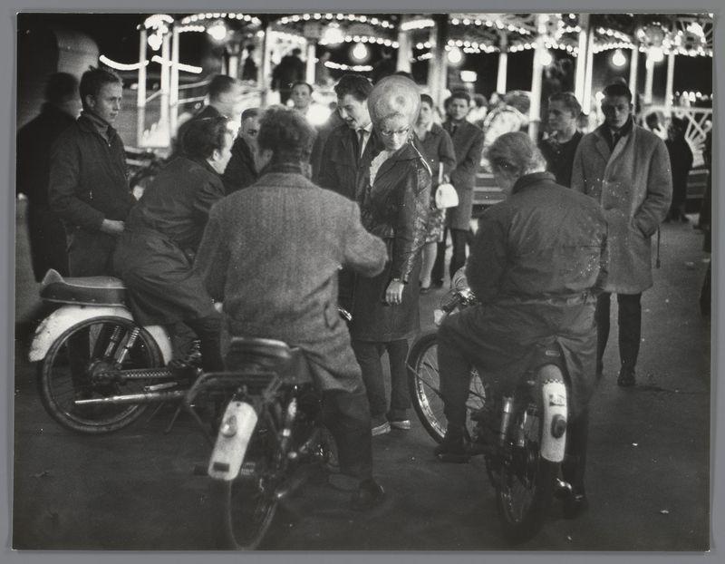 The image depicts a group of people gathered at what appears to be a festive outdoor event, likely in the evening given the illuminated background and ambient lighting. The setting suggests an amusement park or fairground atmosphere with bright lights and structures that resemble rides or decorations typically found at such venues.

In the foreground, there are three individuals on motorcycles. One person is seated directly behind another who stands up from their motorcycle seat to engage with a woman in motion. This interaction seems casual as no formal attire is present; rather, everyone appears dressed for leisurely outdoor activity. The clothing of all subjects looks appropriate for cooler weather.

The background shows more people milling about and engaging in conversation or looking around at the fairground attractions. There's a mix of standing individuals conversing with each other and others who seem to be waiting their turn on rides or simply enjoying the ambiance.

Not